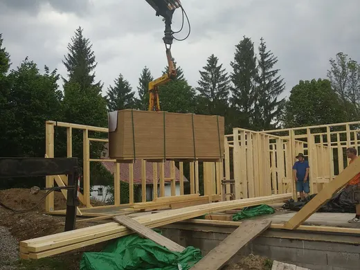 Hydraulická ruka zvedá paletu desek nad rámovou konstrukci dřevostavby